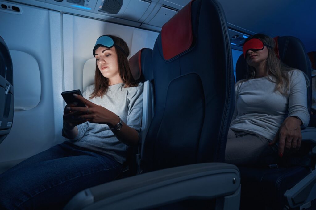 Woman with eye mask on head looking at mobile phone display while flying on plane at night