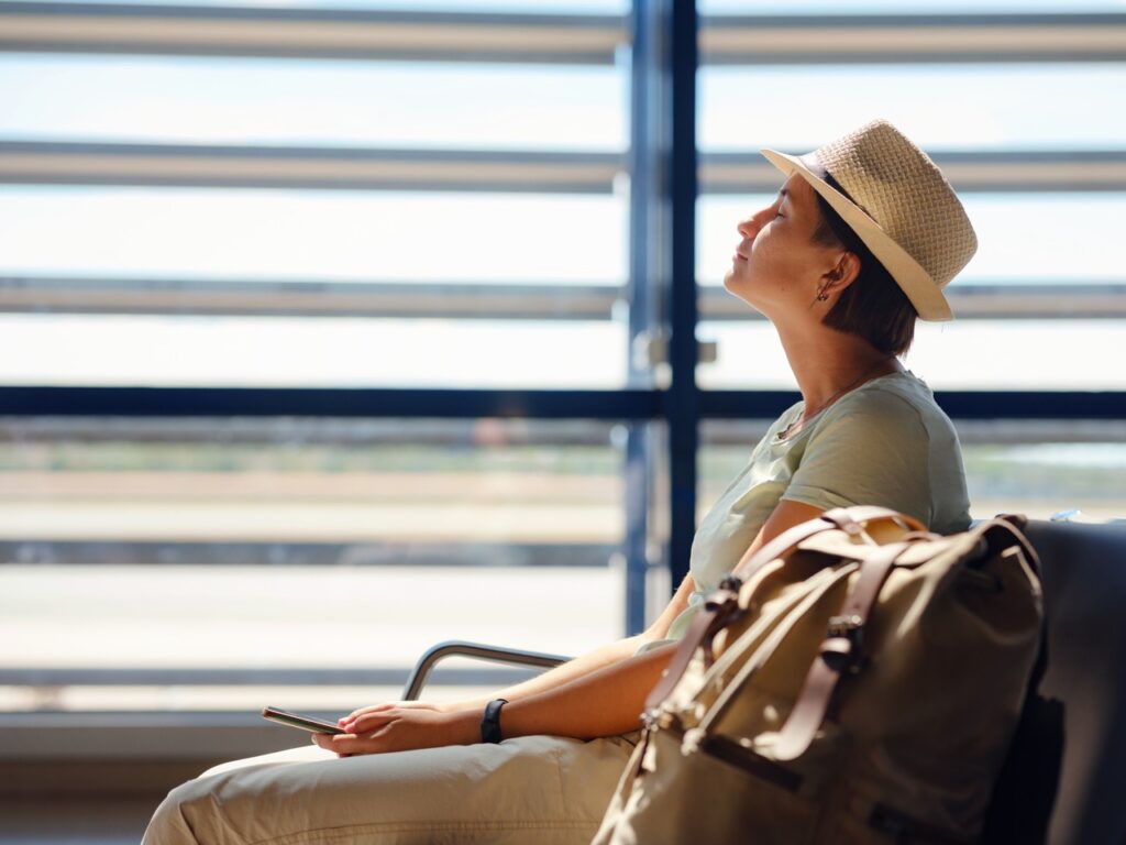 Travel Sentry | ¿A qué hora deberías llegar al aeropuerto? Una joven viajera asiática con una mochila hipster y una gorra, en el aeropuerto de Viena, esperando la salida de su vuelo. Una mujer que viaja se relaja, medita y se tranquiliza antes de subir al avión. Concepto: miedo a volar.